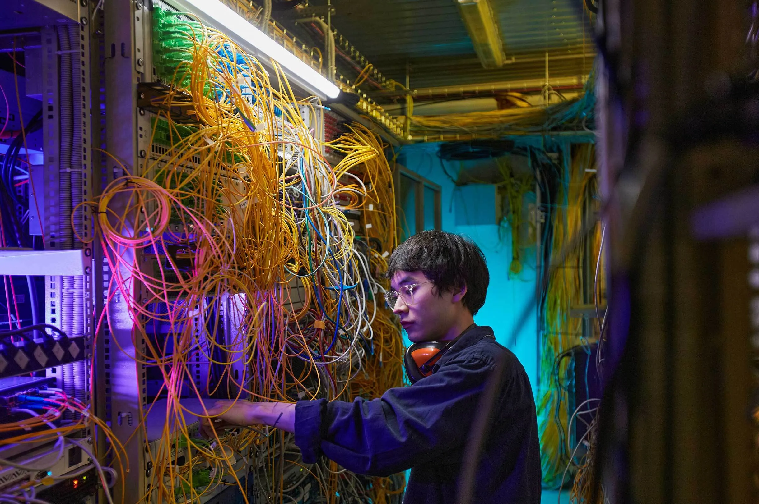 Technician with glasses and headphones adjusting cables in a server room with dense orange and yellow wires.