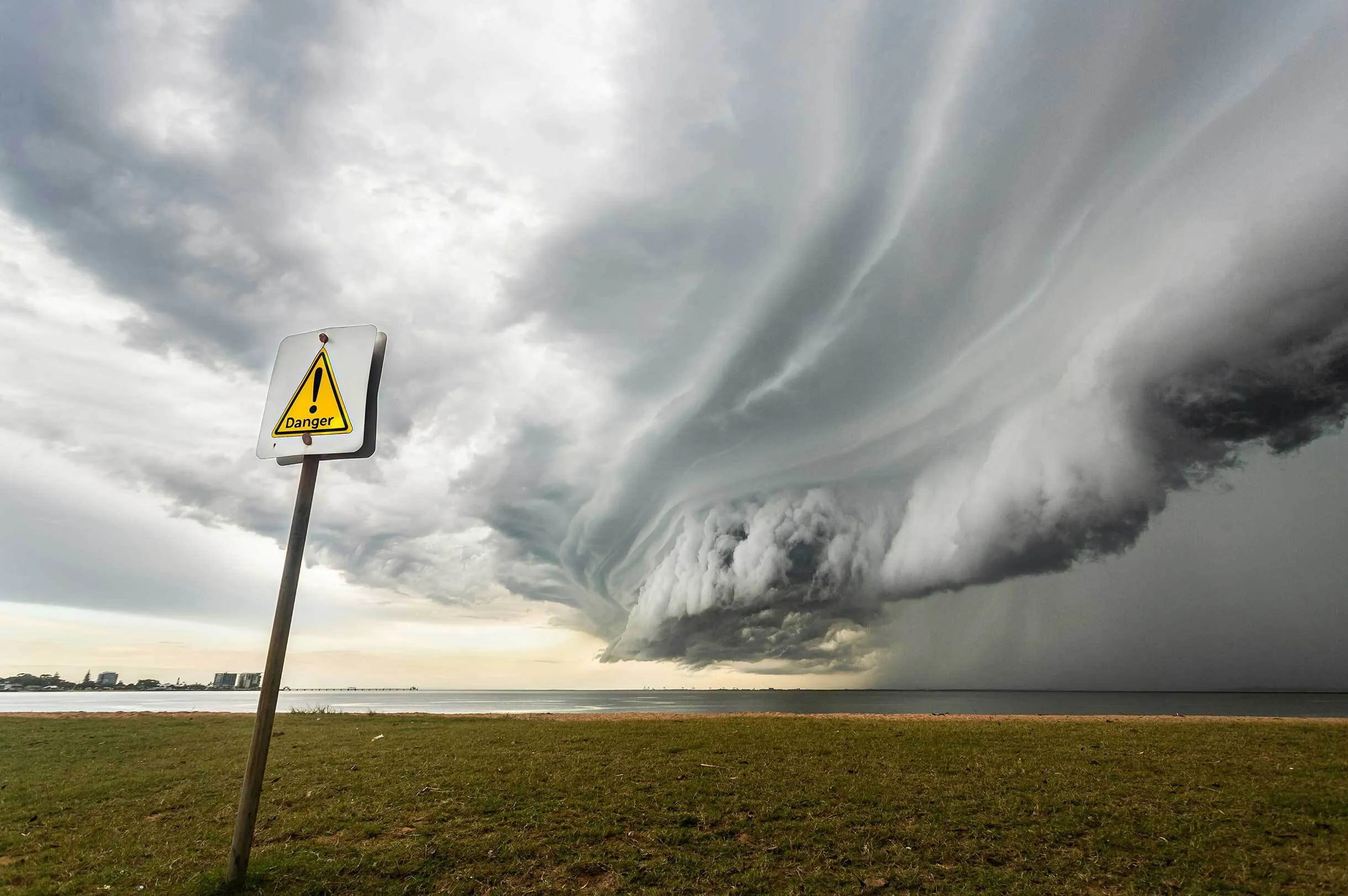 Dramatic dark storm clouds rolling over a body of water with a tilted yellow triangular danger sign in the foreground on grassy land.