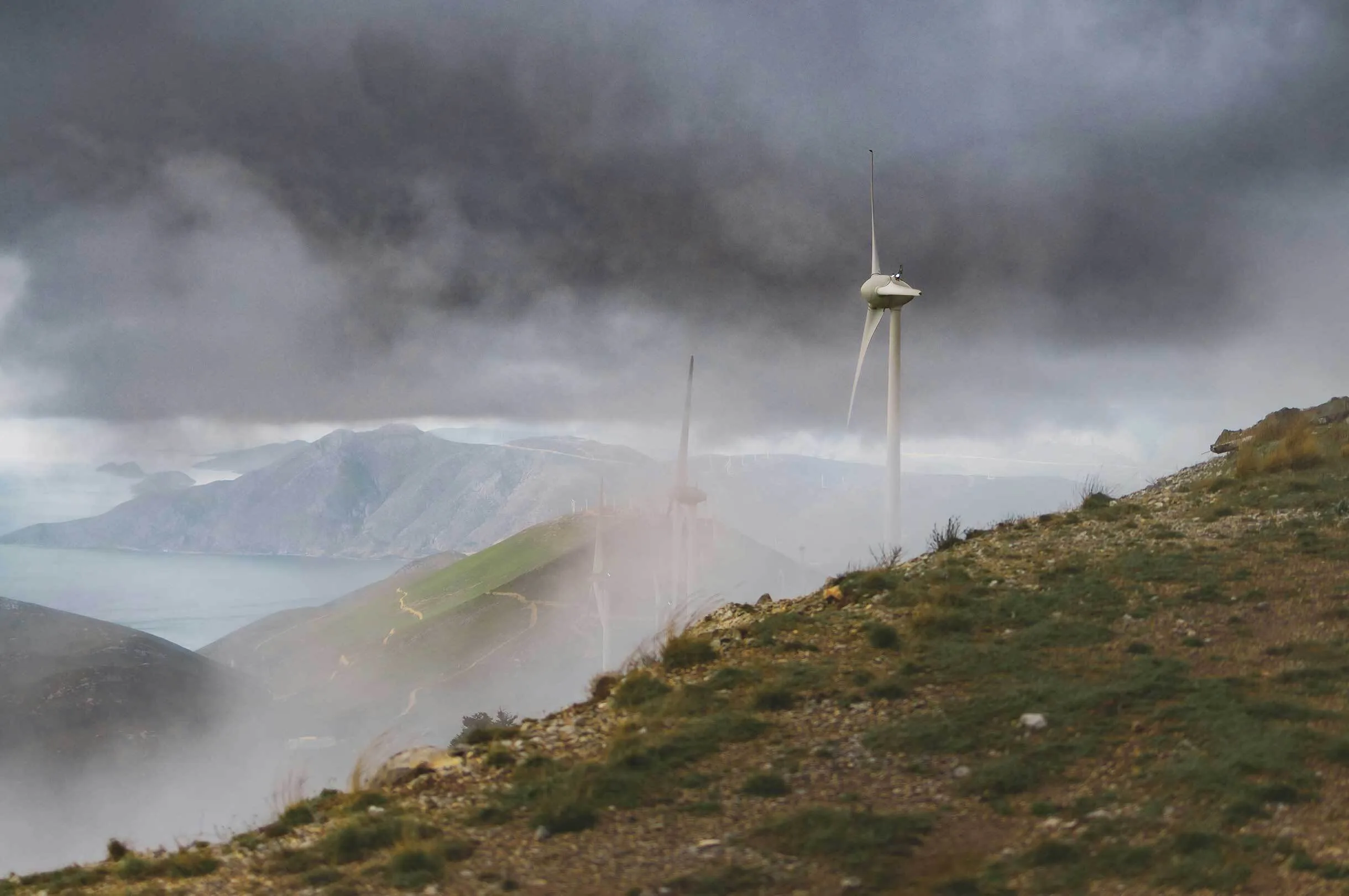 Wind turbines on misty mountain ridge under dark cloudy sky overlooking distant hills and sea.